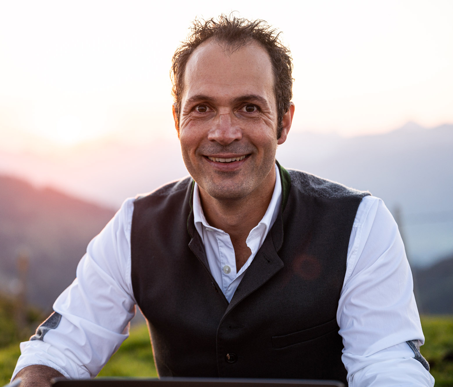 Portraitaufnahme von Johannes Stehle, lächelnd an einem Tisch in den Bergen sitzend, bei angenehmer Sonneneinstrahlung